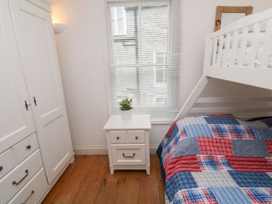 A bedroom with a white wardrobe a white bedside table with a flower pot a bunk bed with a patterned quilt and a window with blinds at Labour in Vain in St Ives
