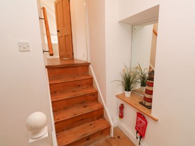 Wooden stairs leading to a door with a handrail a potted plant and lighthouse decoration on a shelf with a mirror above a fire extinguisher and fire blanket on the wall at Labour in Vain in St Ives