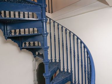 A blue metal spiral staircase with decorative steps and railing against a white wall at Labour in Vain in St Ives