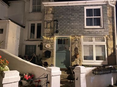 Exterior view of a stone and white house with a green door and windows with string lights above the door at Labour in Vain in St Ives