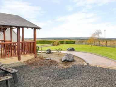 A patio area with a wooden gazebo and gravel ground overlooking a grass lawn and countryside at Alken Cabin in Bushmills County Antrim