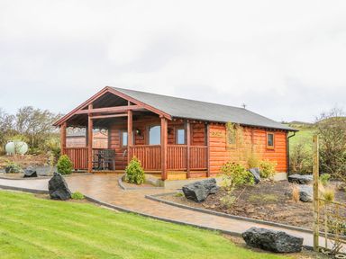 A wooden cabin with a covered porch and a paved path surrounded by grass and rocks at Alken Cabin in Bushmills County Antrim