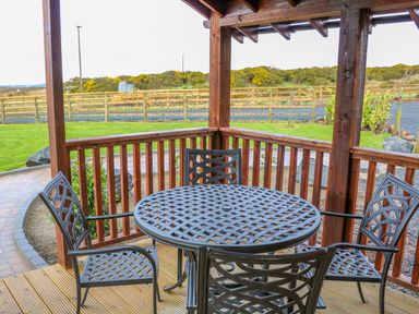 A wooden porch with a round metal table and four chairs overlooking a grassy yard and wooden fence at Alken Cabin in Bushmills County Antrim