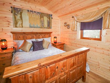 A bedroom with wooden bed frame silver bedding two bedside tables with lamps a window with curtain and blind and artwork on wooden walls at Alken Cabin in Bushmills County Antrim