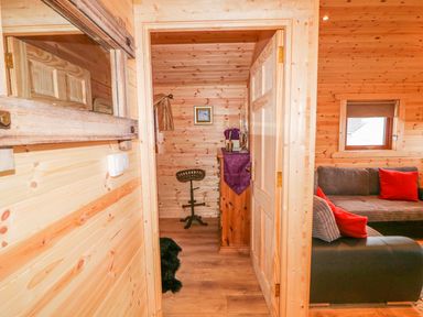 A hallway with wooden walls leading to a small room with a chair and dresser with purple cloth and a living room with a sofa and red pillows at Alken Cabin Bushmills County Antrim
