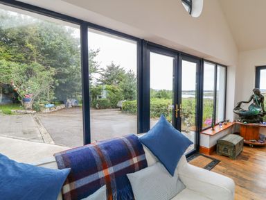 A living room with a sofa and cushions near large windows overlooking a garden and parking area at The Mermaid in Dungloe County Donegal