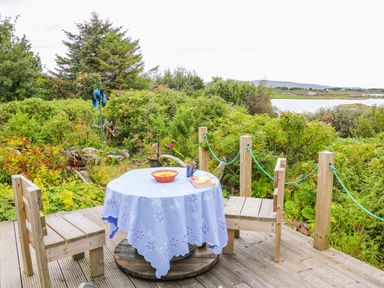 A wooden deck with a round table covered by a blue tablecloth and two wooden chairs surrounded by shrubs and a lake in the background at The Mermaid in Dungloe County Donegal