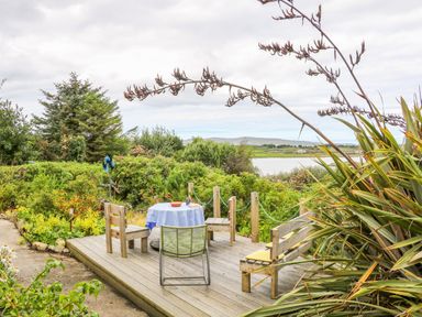 An outdoor wooden deck with chairs and a table covered with a tablecloth overlooking a garden and a body of water at The Mermaid in Dungloe County Donegal