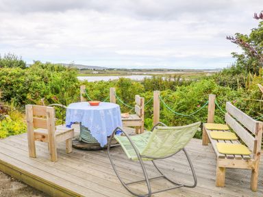 An outdoor wooden deck with chairs a table and a bench overlooking greenery and water at The Mermaid in Dungloe County Donegal