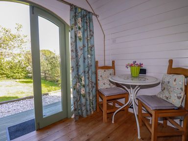 A small dining area with two wooden chairs and a round table next to a glass door with floral curtains at The Lamb Shack in St Teath