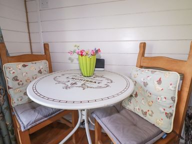 A small round table with a green vase of flowers and two wooden chairs with cushions at The Lamb Shack in St Teath