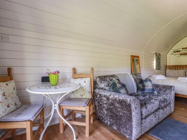 A small sitting area with a round table and two wooden chairs with cushions next to a velvet sofa and a bed in a room at The Lamb Shack in St Teath