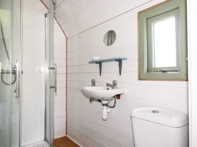 A bathroom with a shower cubicle sink with two taps a small round mirror a shelf and a window at The Lamb Shack in St Teath