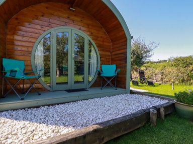 An outdoor wooden pod with a round glass door flanked by two blue folding chairs and white pebbles in front at The Lamb Shack in St Teath