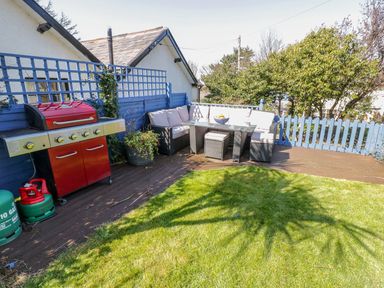 An outdoor patio with a red barbecue grill a cushioned corner sofa glass table potted plants and grass at Penholm in Penmaenmawr
