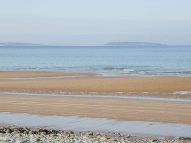 A sandy beach with shallow water and stones in the foreground and an island in the distance at Penholm in Penmaenmawr