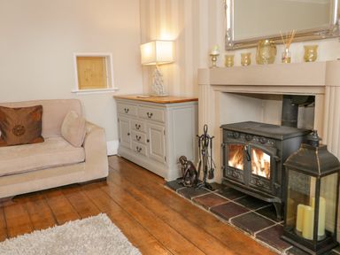 A living room with a beige sofa a gray cabinet with a lamp a black wood stove fire a mirror and a large lantern at Penholm in Penmaenmawr