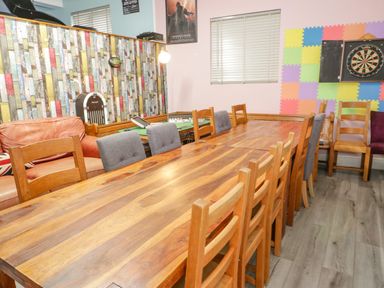 A dining room with a long wooden table surrounded by wooden and cushioned chairs with a dartboard and colorful foam wall panels at Penholm in Penmaenmawr