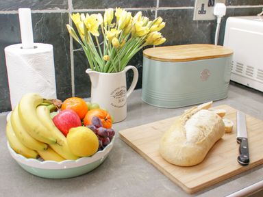 A kitchen countertop with a bowl of fruit a loaf of bread with slices on a cutting board a knife a bread box a roll of paper towels and a jug with yellow flowers at Penholm in Penmaenmawr