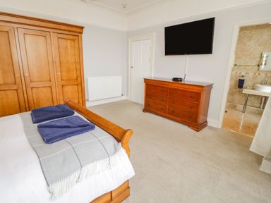 A bedroom with a wooden bed with towels on it wooden wardrobe wooden dresser and wall mounted TV at Penholm Penmaenmawr