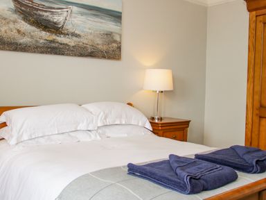 A bedroom with a wooden bed with white linens folded navy towels bedside table with a lamp and a wooden wardrobe at Penholm in Penmaenmawr