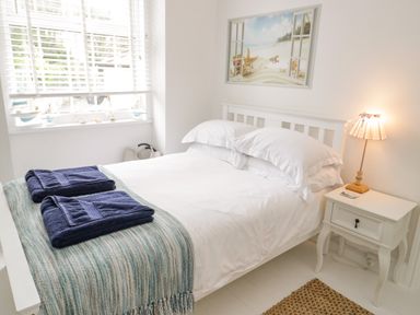 A bedroom with a white bed and pillows a light blue throw with two navy towels on top a white nightstand with a lamp and a window with blinds and decorative boats at Penholm Penmaenmawr