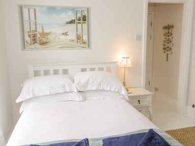 A bedroom with a white bed frame white bedding a bedside table with a lamp and a beach-themed wall art at Penholm in Penmaenmawr