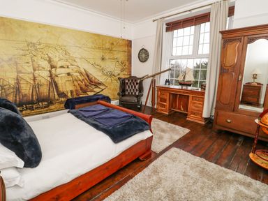 A bedroom with a wooden bed, a leather chair, a wooden desk with a model ship, and a wooden wardrobe at Penholm in Penmaenmawr