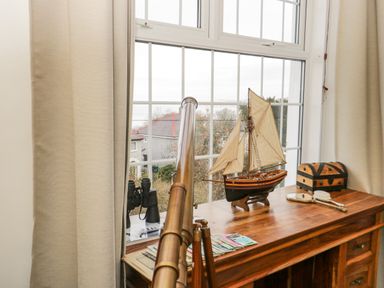 A wooden table with a model ship telescope binoculars and a wooden chest by a window with beige curtains at Penholm Penmaenmawr
