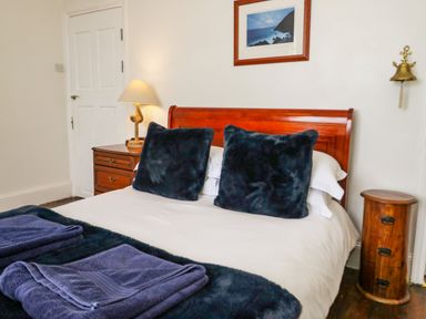 A bedroom with a bed with black pillows and towels a wooden bedside table with a lamp a round wooden cabinet a framed picture and a wall-mounted bell at Penholm in Penmaenmawr