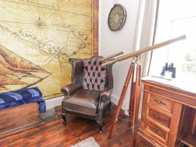 A leather armchair with a brass telescope on a tripod next to a wooden chest and a wooden cabinet with binoculars in a room with a large vintage map on the wall at Penholm in Penmaenmawr