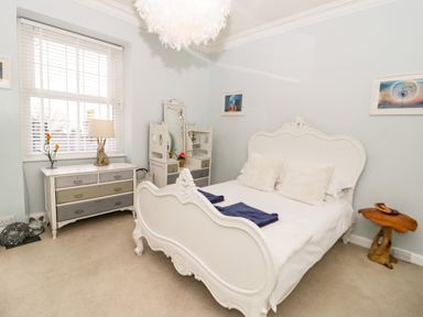 A bedroom with a white ornate bed two blue towels on bed white dresser with lamp and flowers window with blinds at Penholm in Penmaenmawr