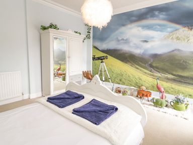 A bedroom with a white bed and blue towels a wardrobe with a mirror a telescope and a wall mural of a mountain landscape with a rainbow at Penholm in Penmaenmawr