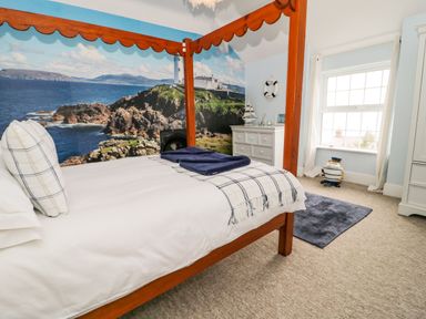 A bedroom with a four poster bed a dresser a window and a large wall mural of a coastal lighthouse at Penholm Penmaenmawr