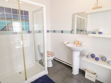 A bathroom with a shower, toilet, pedestal sink, shelves with decorative items, and tiled walls at Penholm in Penmaenmawr