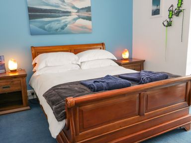 A wooden bed with white bedding and blue towels on a blanket between two wooden nightstands with lamps and a blue wall with a landscape picture at Penholm in Penmaenmawr