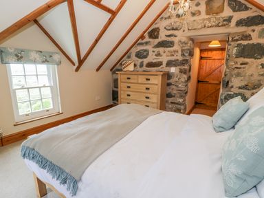 A bedroom with a bed with cushions and a throw blanket a wooden chest of drawers a window and a stone wall with a wooden door at Llety Perygl in Harlech