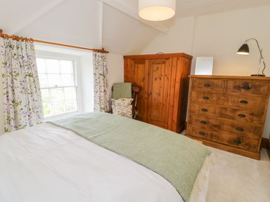 A bedroom with a bed a wooden wardrobe a wooden dresser a chair with cushions and floral curtains at Llety Perygl Harlech