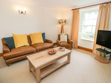 A living room with a brown leather sofa and armchair a wooden coffee table a floor lamp a television and a window with beige curtains at Llety Perygl in Harlech