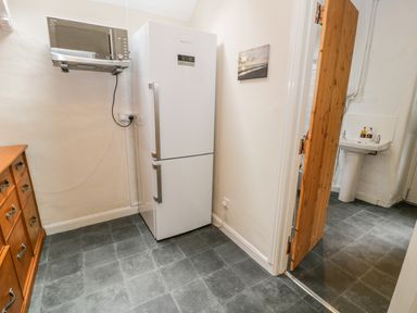 A kitchen area with a white refrigerator, a microwave mounted on the wall, a wooden cabinet with drawers, and a doorway leading to a room with a sink at Llety Perygl in Harlech
