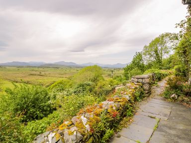 A stone pathway bordered by a moss-covered stone wall with plants and trees overlooking a green field and distant mountains at Llety Perygl in Harlech