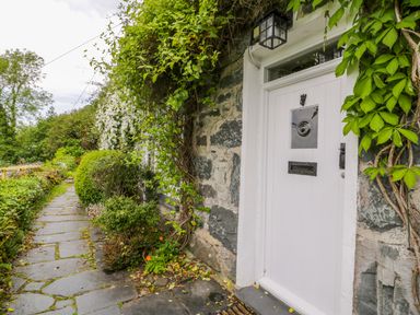 A stone path with plants and greenery leading to a white door on a stone building at Llety Perygl in Harlech