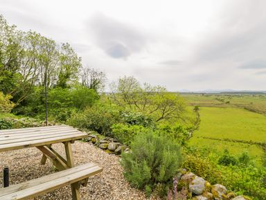 An outdoor picnic table on a gravel area with green bushes and trees overlooking a grassy field and distant hills at Llety Perygl in Harlech
