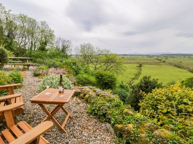 A garden with wooden chairs and a table with a bottle and glasses overlooking green fields at Llety Perygl in Harlech