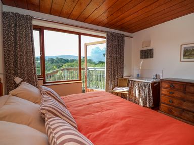 A bedroom with a bed covered in an orange duvet a wooden chest of drawers a small sink and chair next to a large window with curtains at Plasybangor in Newport Pembrokeshire