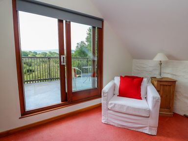 A room with a white armchair with a red pillow next to a wooden side table with a lamp near a glass door leading to a balcony at Plasybangor in Newport Pembrokeshire