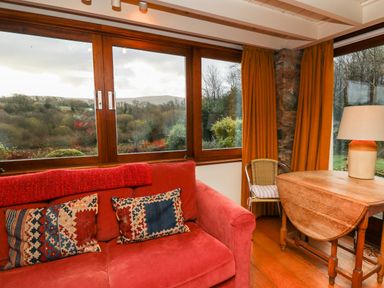 A room with a red sofa with patterned cushions a wooden table with a lamp and a chair near large windows with mustard curtains at Plasybangor Newport Pembrokeshire