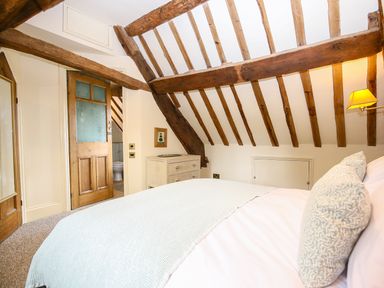 A bedroom with exposed wooden beams a bed with pillows a dresser a wall lamp and an open door showing a bathroom at Murton Cottage in Burford