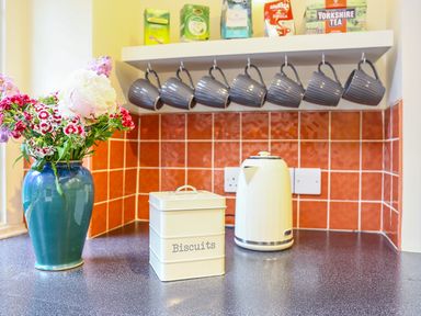 A kitchen corner with a vase of flowers a biscuit tin a kettle hanging mugs and tea boxes on a shelf at Murton Cottage in Burford