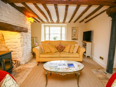 A living room with a yellow sofa a marble coffee table wooden ceiling beams and a stone fireplace at Murton Cottage in Burford